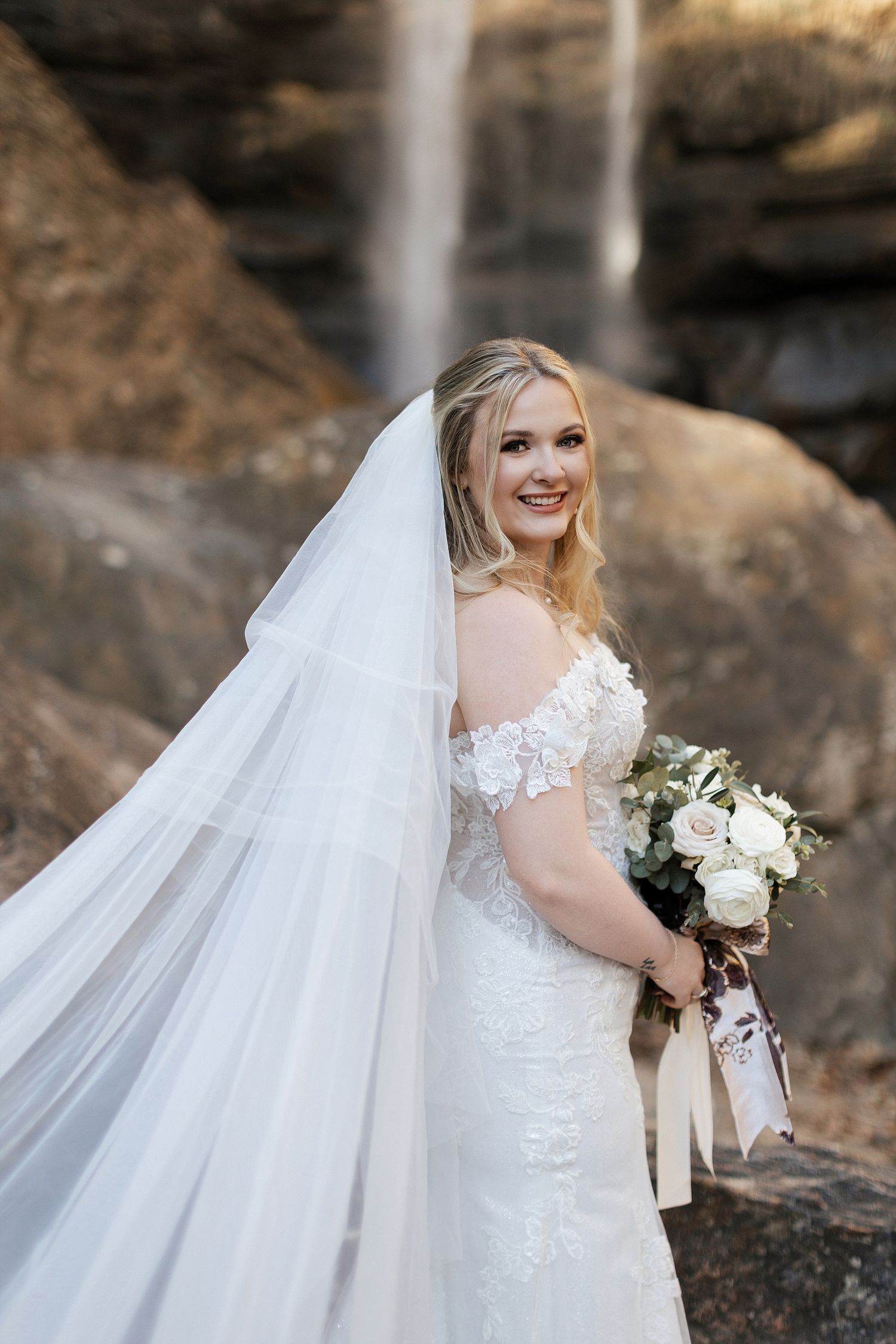 Beautiful bridal portrait with flowing veil and white bouquet captured against scenic waterfall backdrop.