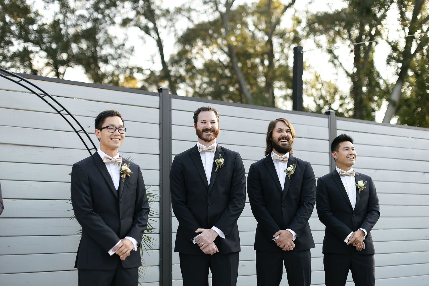 Four groomsmen in black suits and ties standing in front of a white horizontal plank wall.