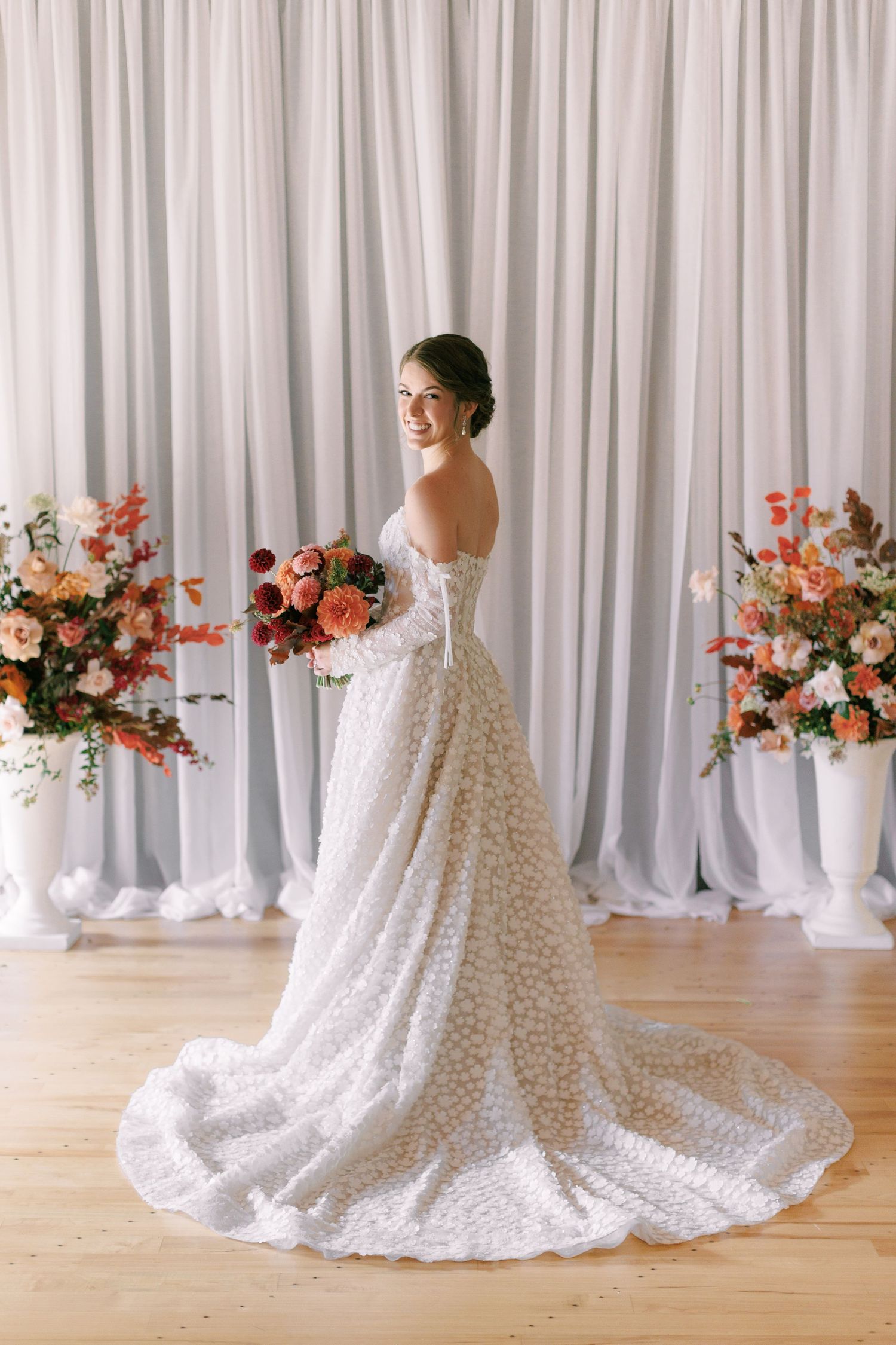 Smiling bride in a floral lace gown with a long train holds a bouquet, posing before white drapes and floral arrangements.