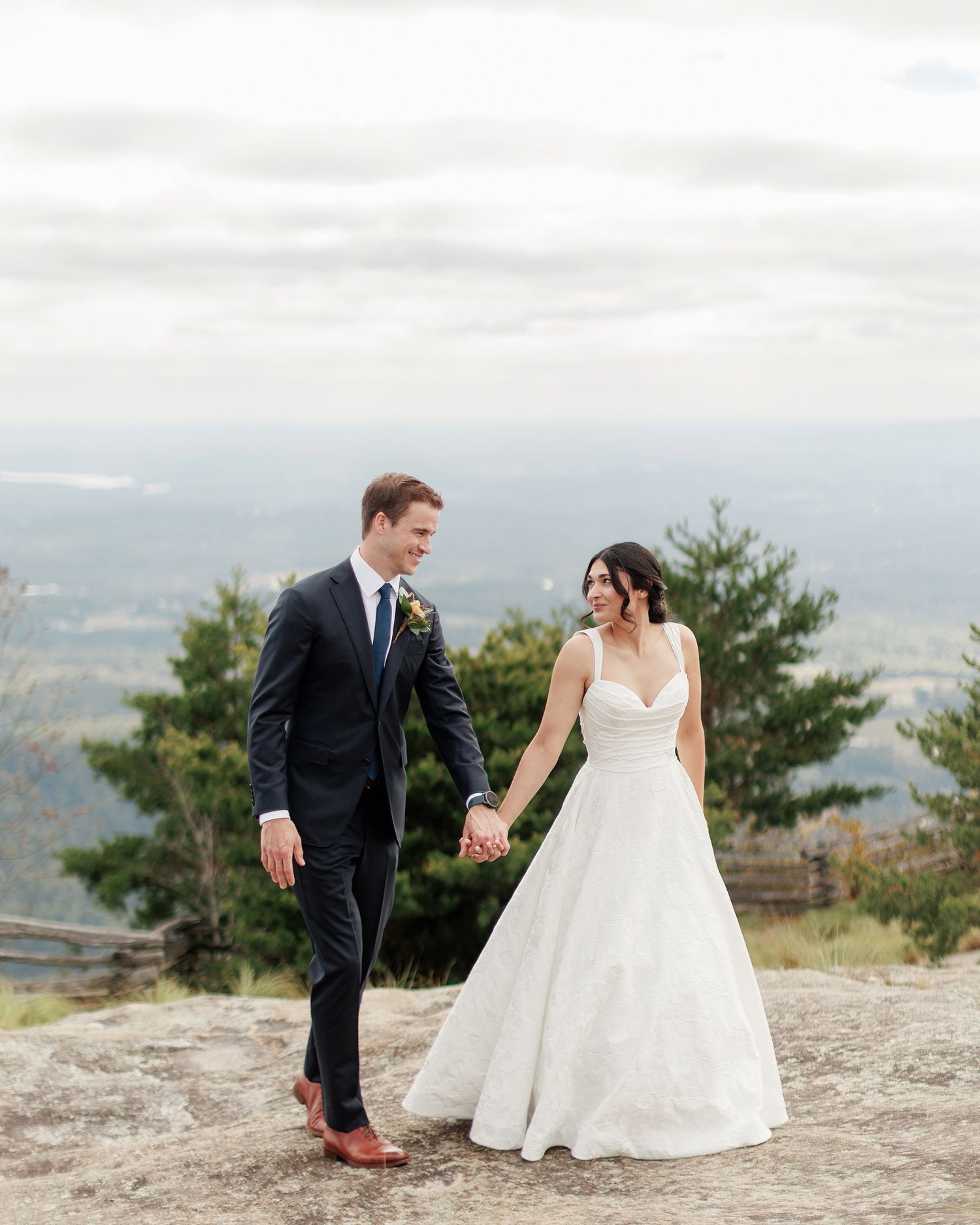 A couple in wedding attire sits together on a rocky cliff overlooking a scenic mountain landscape.