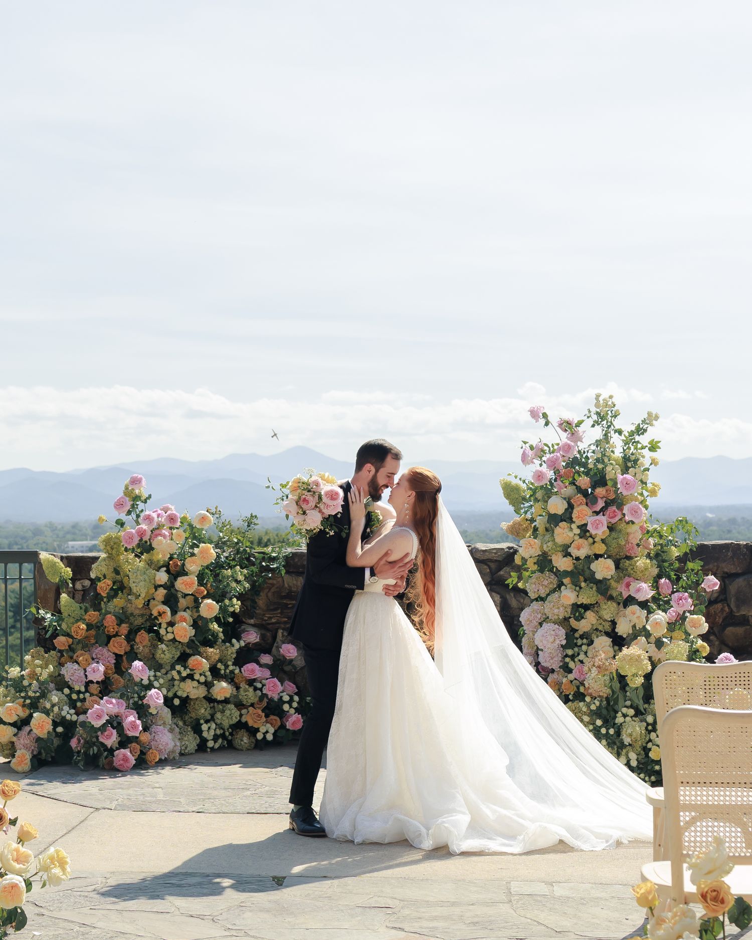 Bride and groom share a romantic kiss at an outdoor wedding ceremony surrounded by lush pink and yellow floral arrangements.
