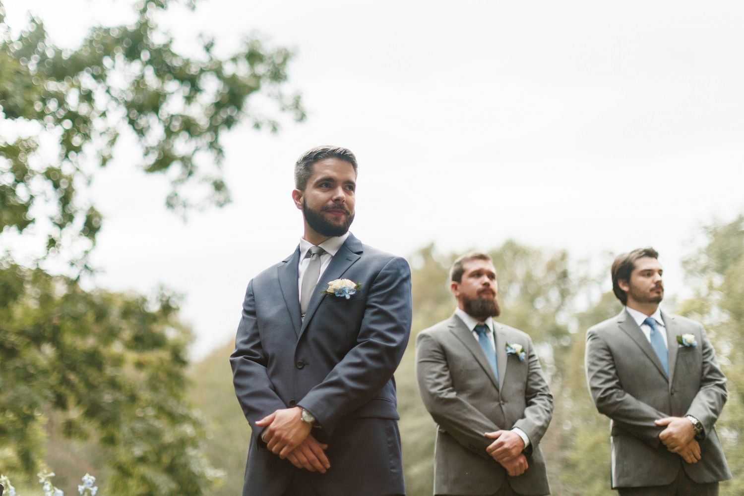 Three groomsmen in gray suits standing outdoors during a wedding ceremony, with trees in the background.