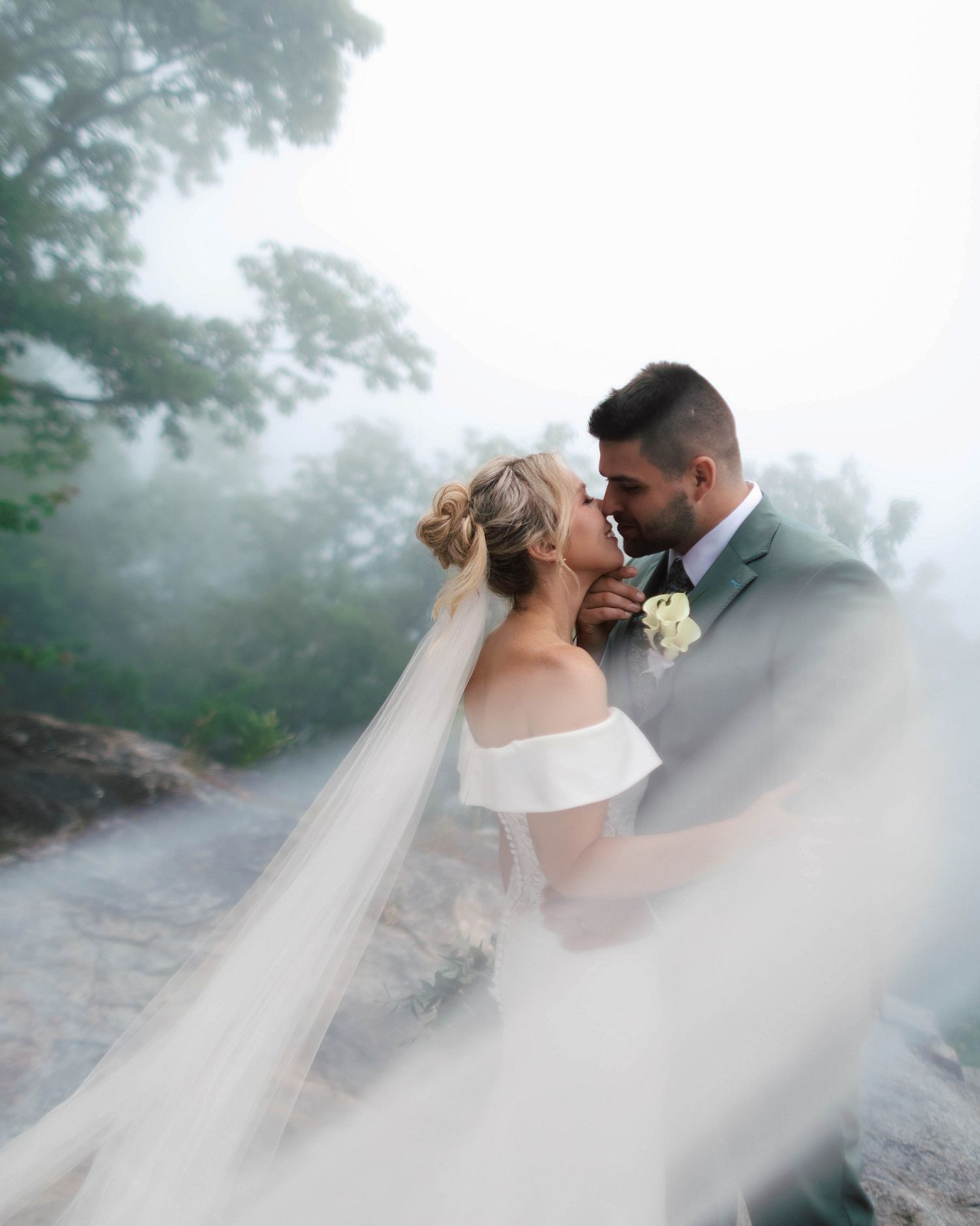 Bride and groom sharing a kiss on a foggy mountain, bride's veil flowing dramatically in the foreground.