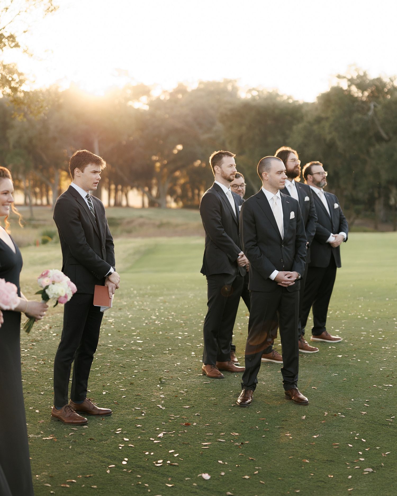 Wedding party in formal attire poses on a grassy field at golden hour.