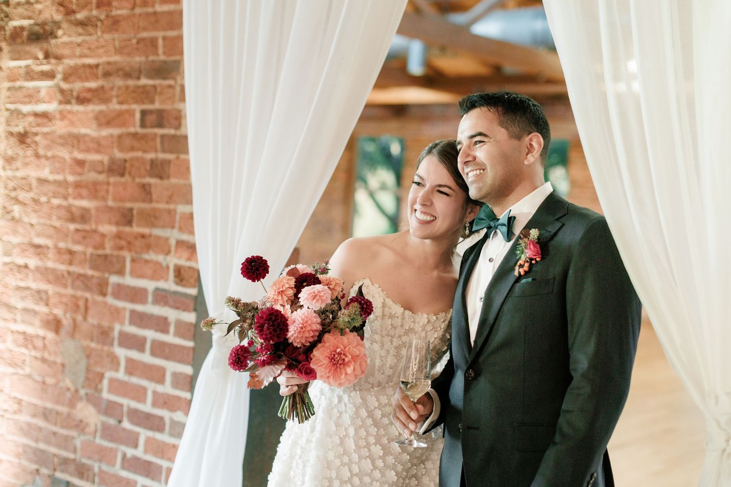 Bride and groom smiling together under white draping near brick wall, bride holding pink and red floral bouquet.