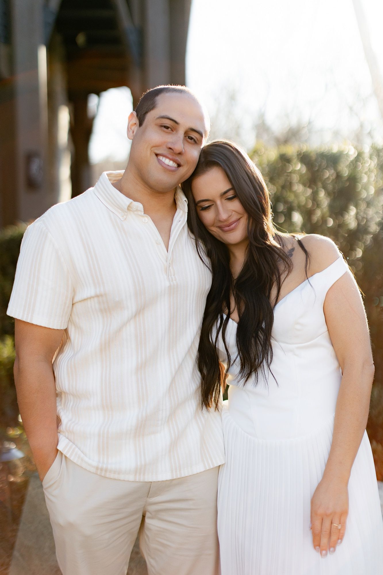 Couple in coordinated white outfits posing together in warm sunlight.