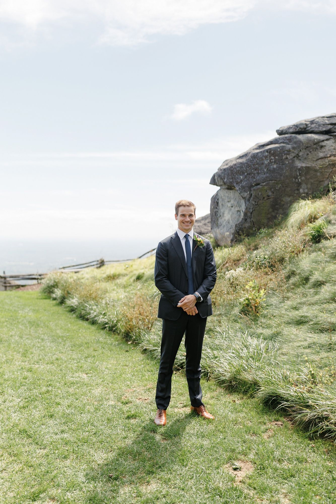 A person in a dark suit poses on a grassy hillside overlooking a scenic coastal landscape.