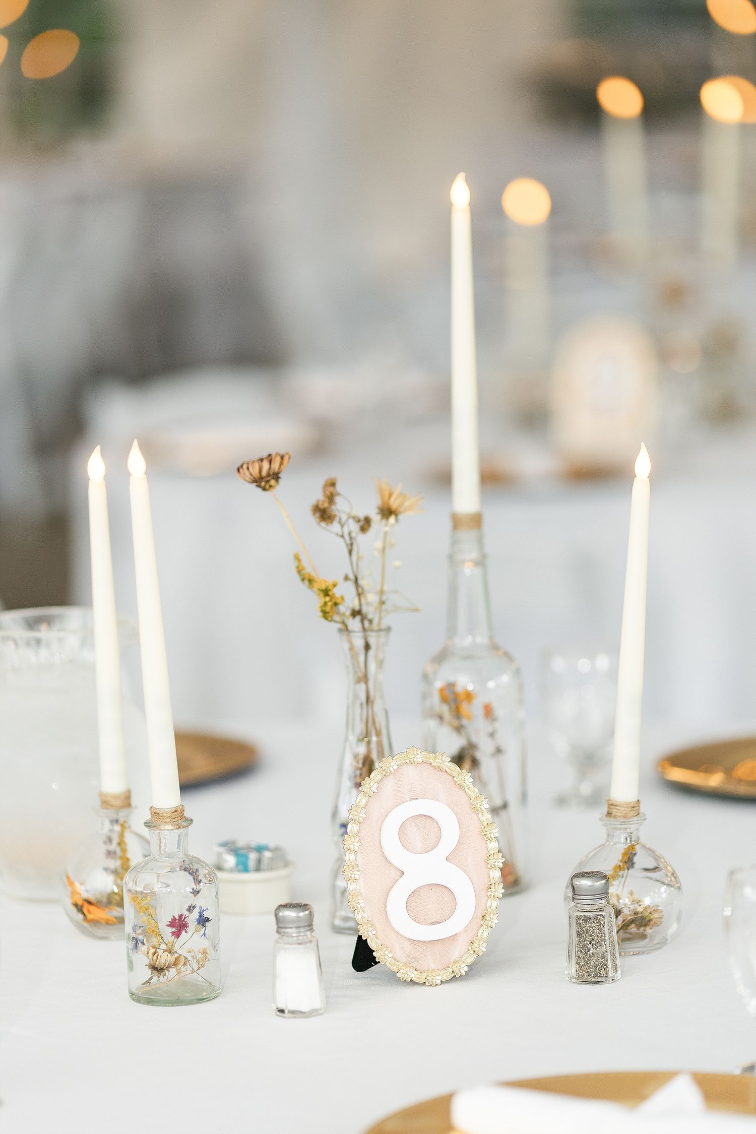 Elegant table centerpiece with white candles, glass bottles, dried flowers and a wooden number 8 table marker.