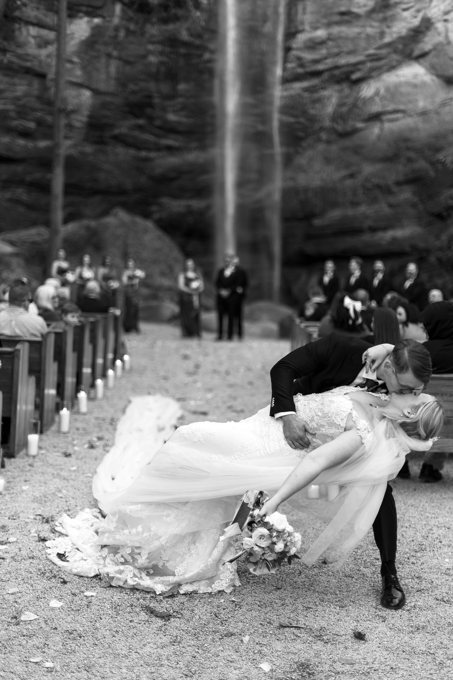 A dramatic black and white wedding photo showing a couple dancing near a waterfall with guests watching in the background.