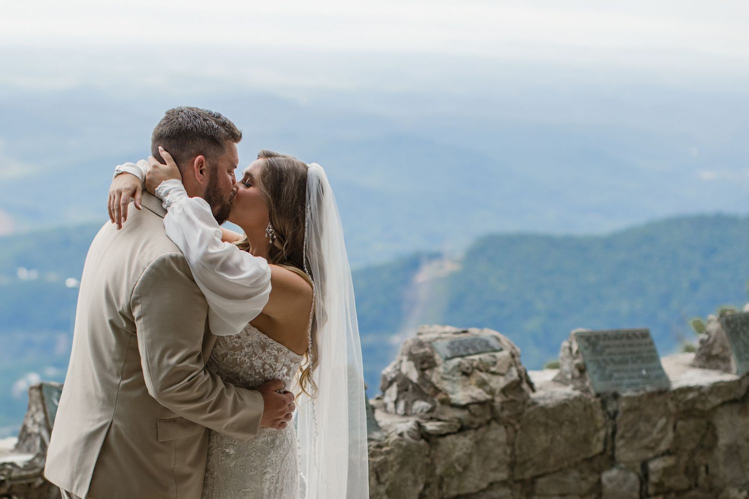 A romantic wedding couple shares a kiss against a backdrop of mountain peaks and ancient stone ruins.