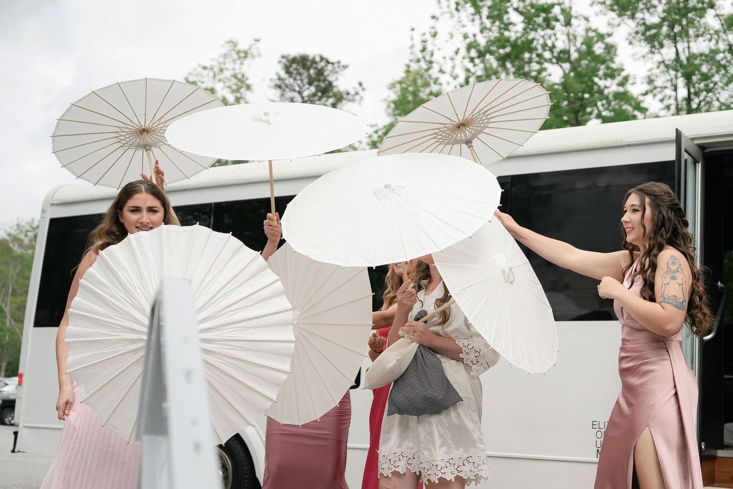 Wedding party members twirl white paper parasols outside while standing next to a white van in an outdoor setting.
