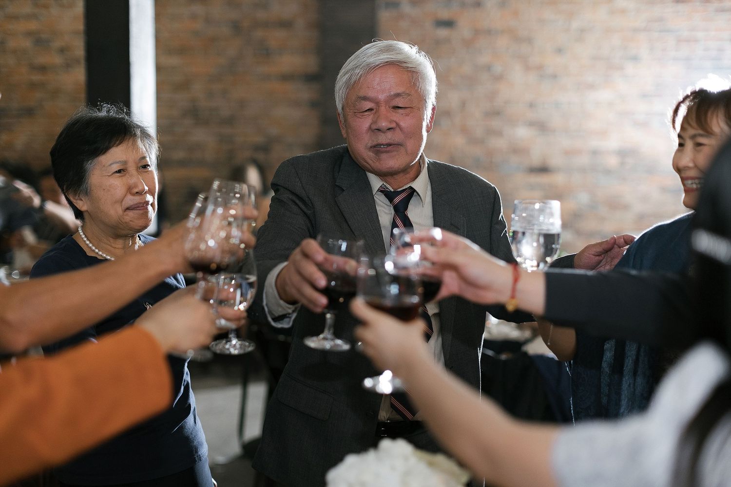 People raise their wine glasses in a toast at a gathering with exposed brick walls in the background.
