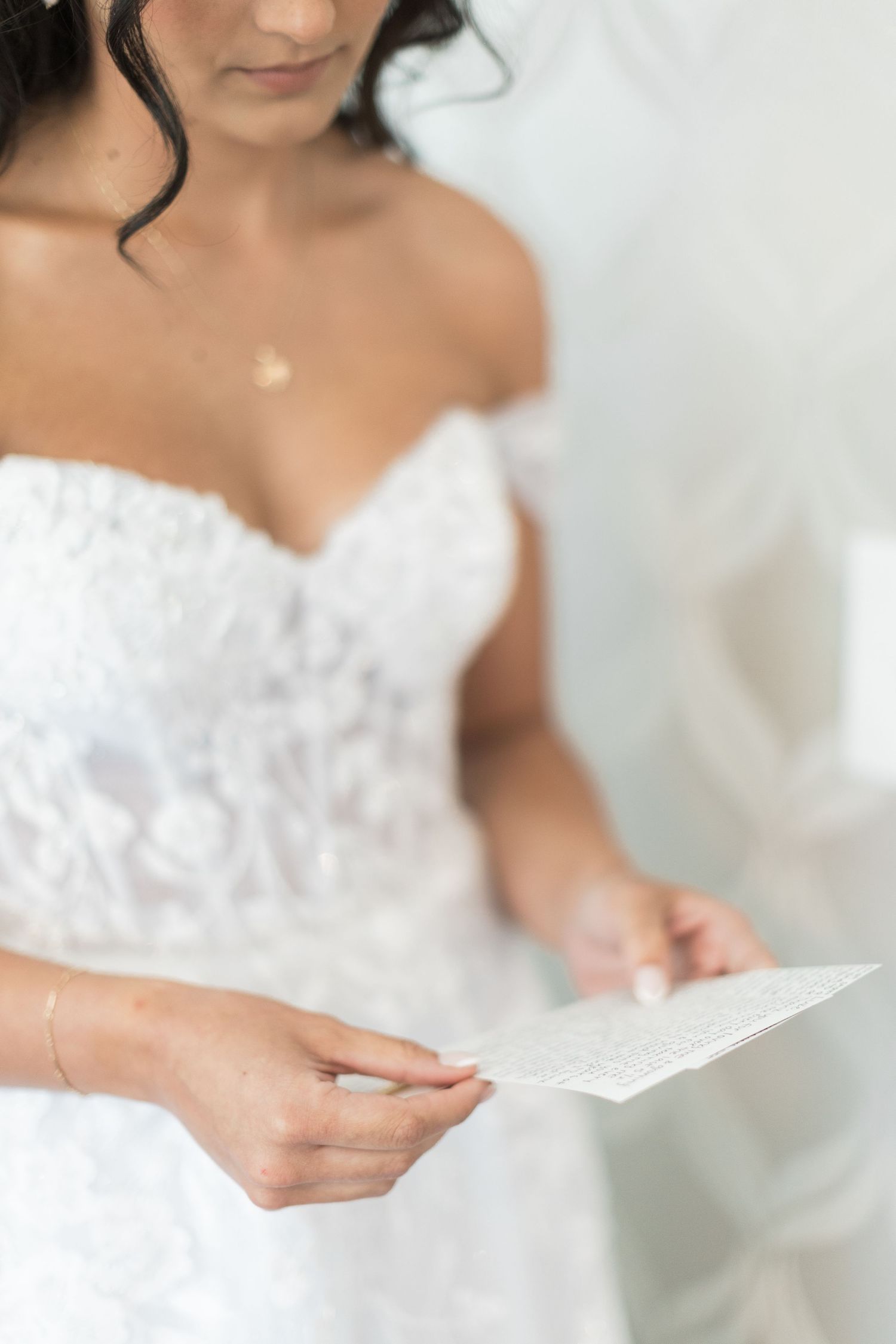 Bride in off-shoulder lace wedding gown reading a heartfelt letter, captured in soft natural light.