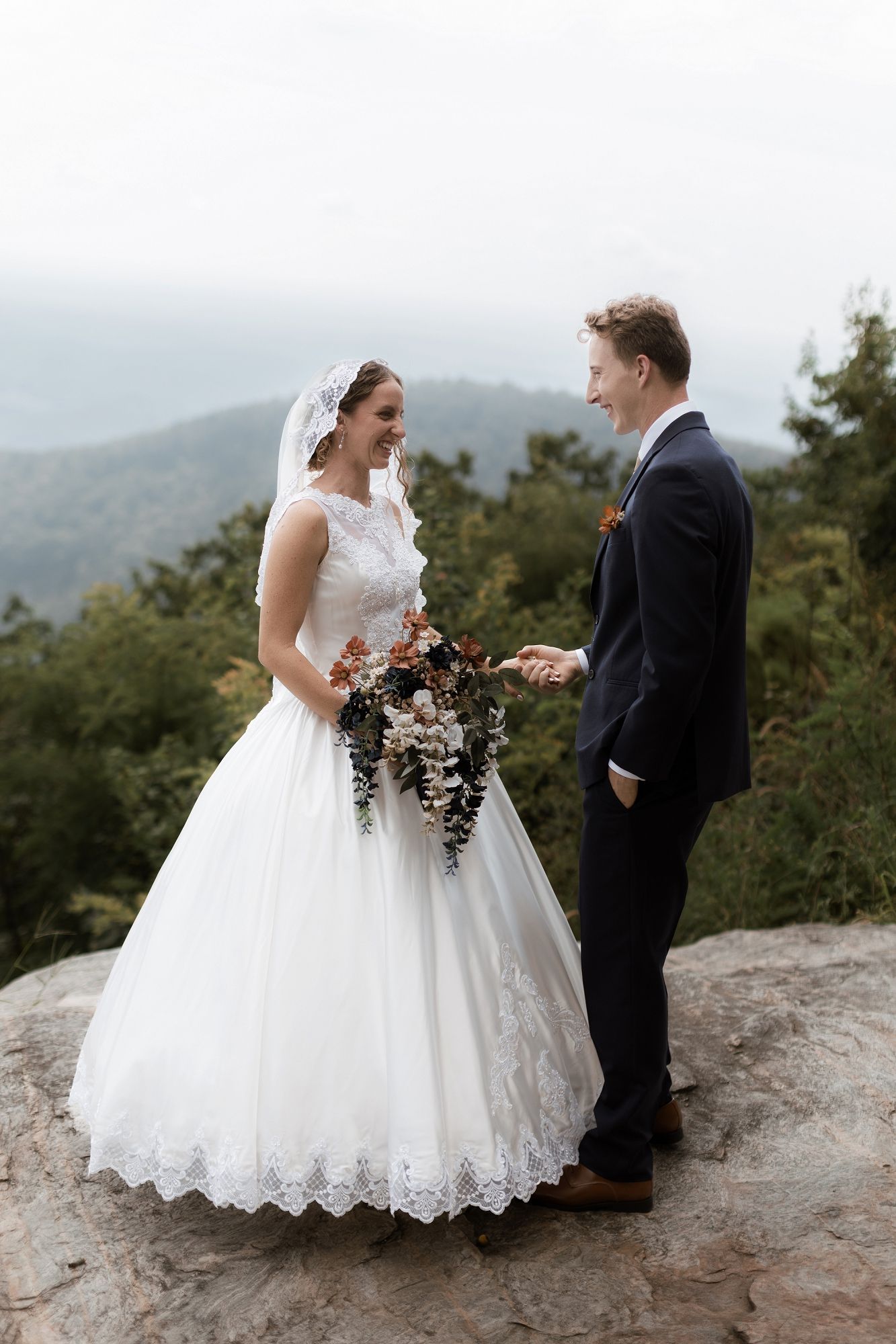 Smiling bride and groom stand on a rocky mountain overlook, bride holding a cascading bouquet.