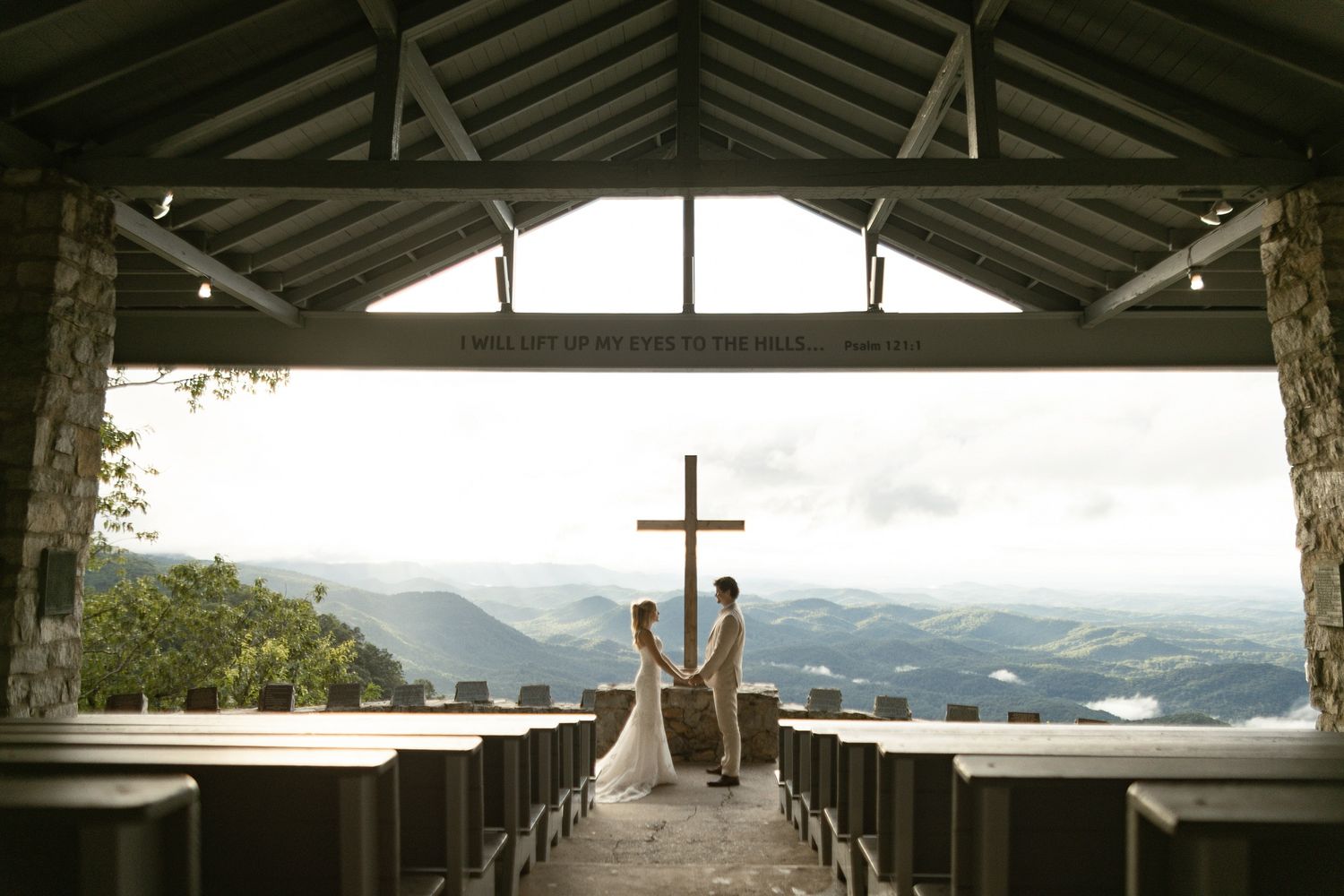 Bride and groom holding hands at altar before wooden cross in open-air mountain chapel.