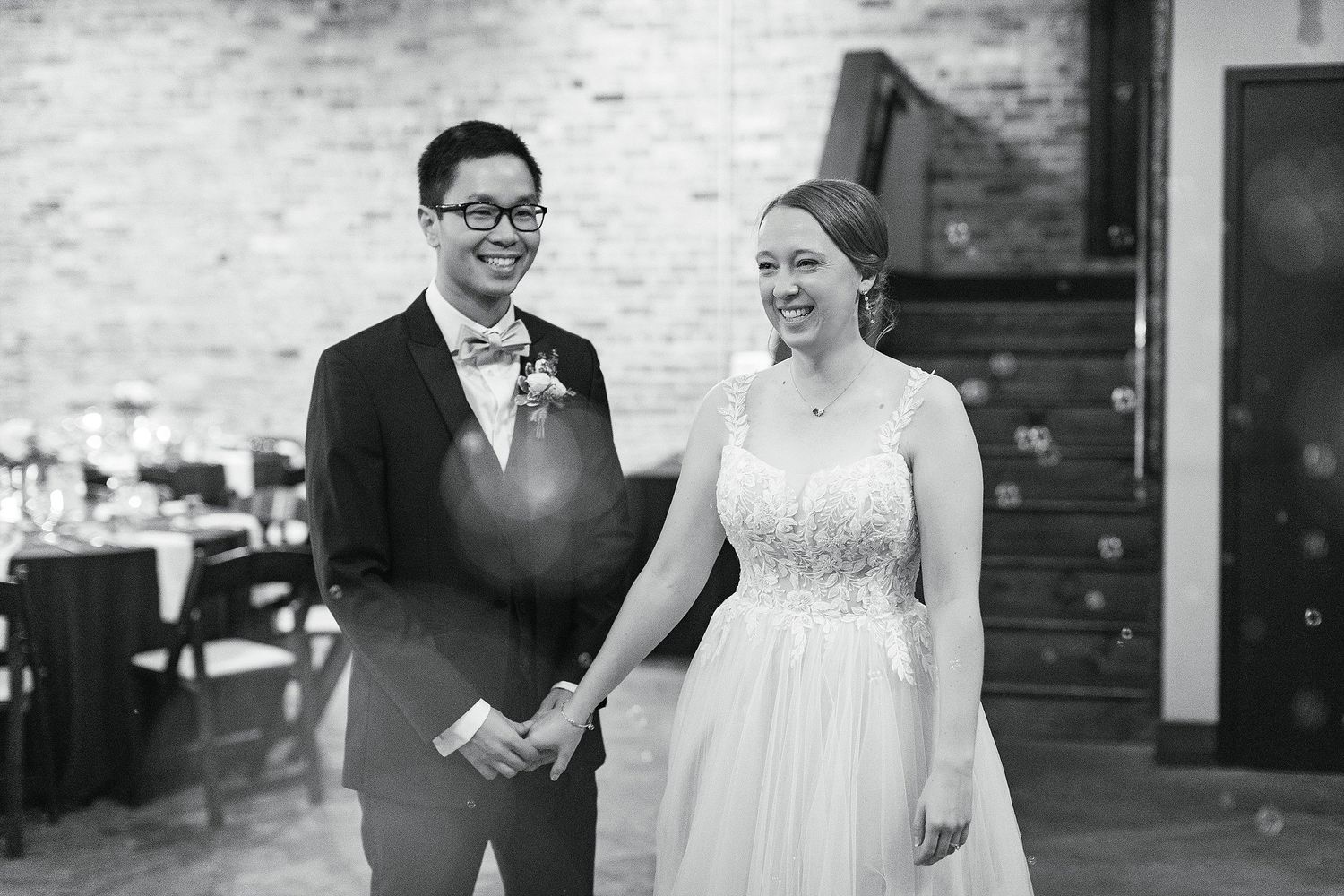 A black and white wedding photo showing a couple laughing together in a rustic brick-walled venue.