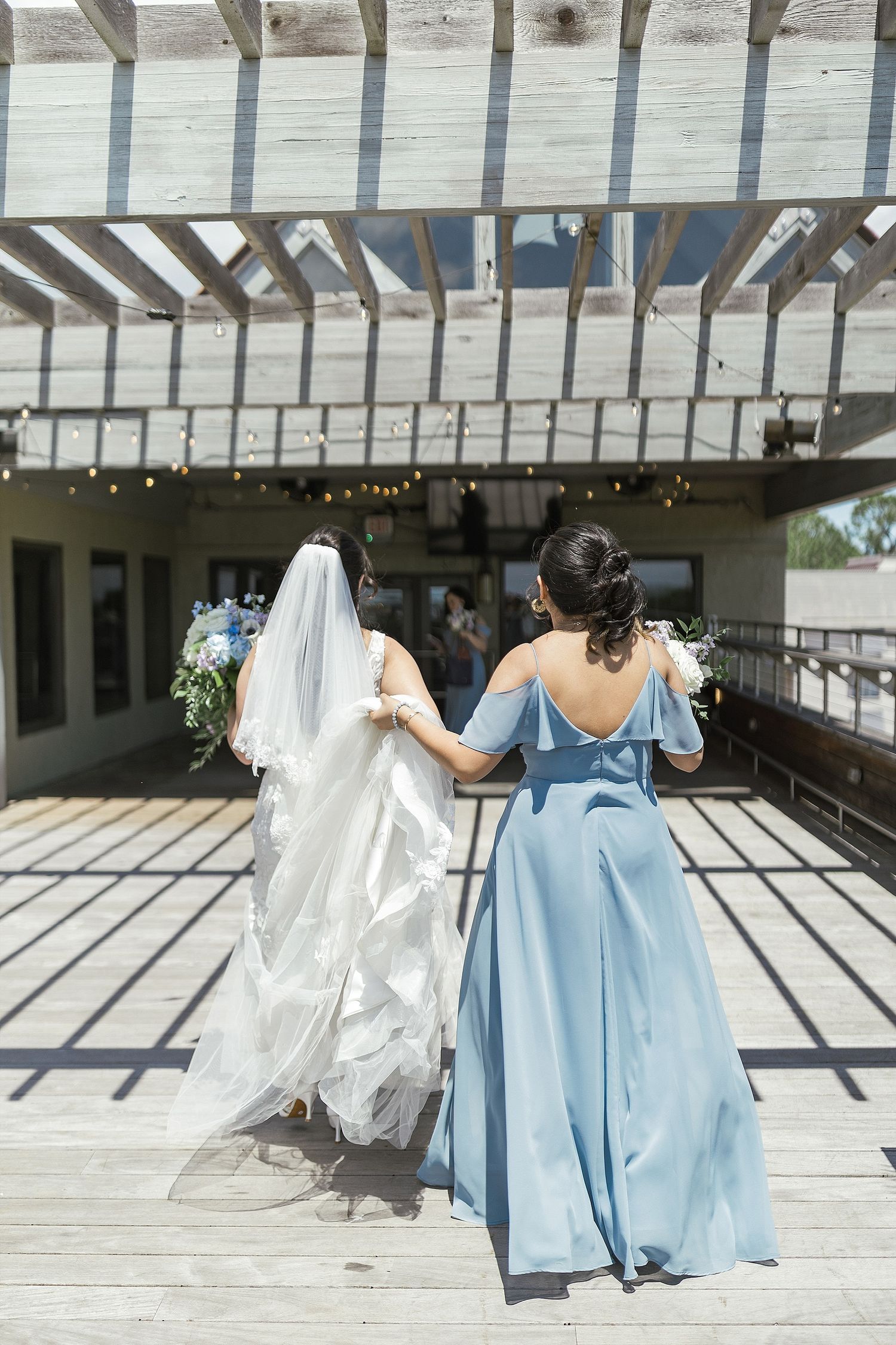Bride and bridesmaid walking under pergola casting dramatic shadows on wooden deck.