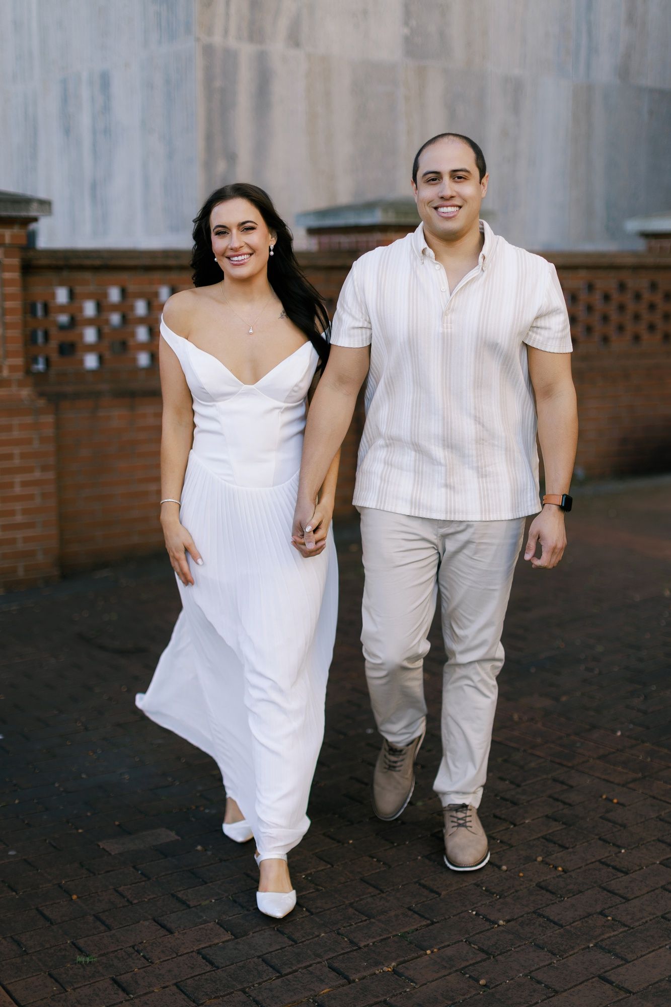 A couple walking together outdoors, with her in a flowing white dress and him in a casual white striped shirt and khakis.