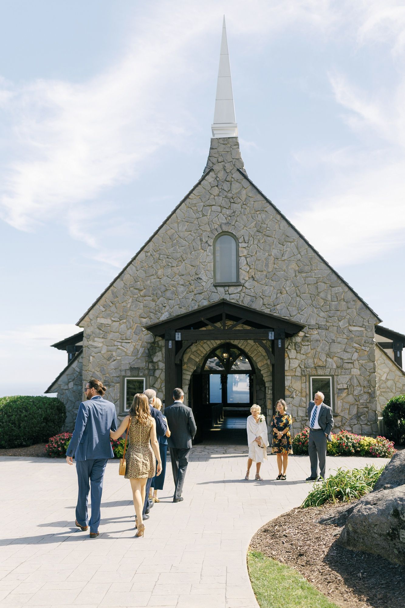Stone chapel exterior with arched entrance and steeple set against blue sky at a scenic mountain location.