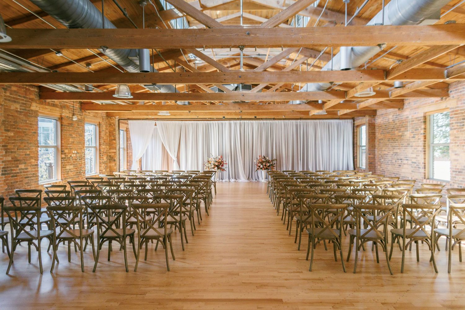 Elegant wedding ceremony venue with exposed brick walls, wooden beam ceiling, crossback chairs, and a draped altar backdrop.