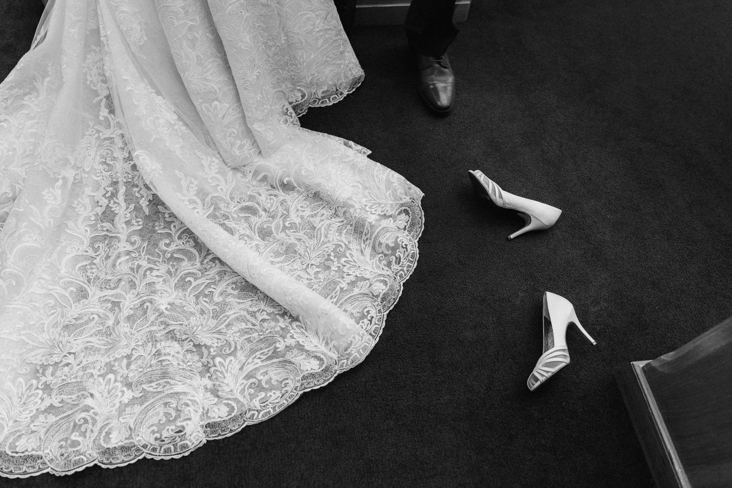 Black and white photo of a lace wedding dress train with white heels on carpet beside it.