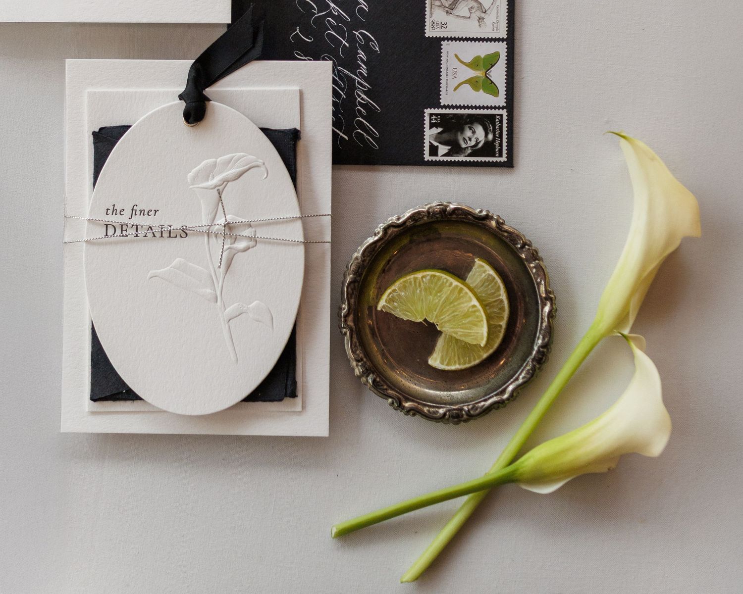 Elegant wedding stationery flatlay with embossed card, calla lilies, and lemon slice on silver dish.