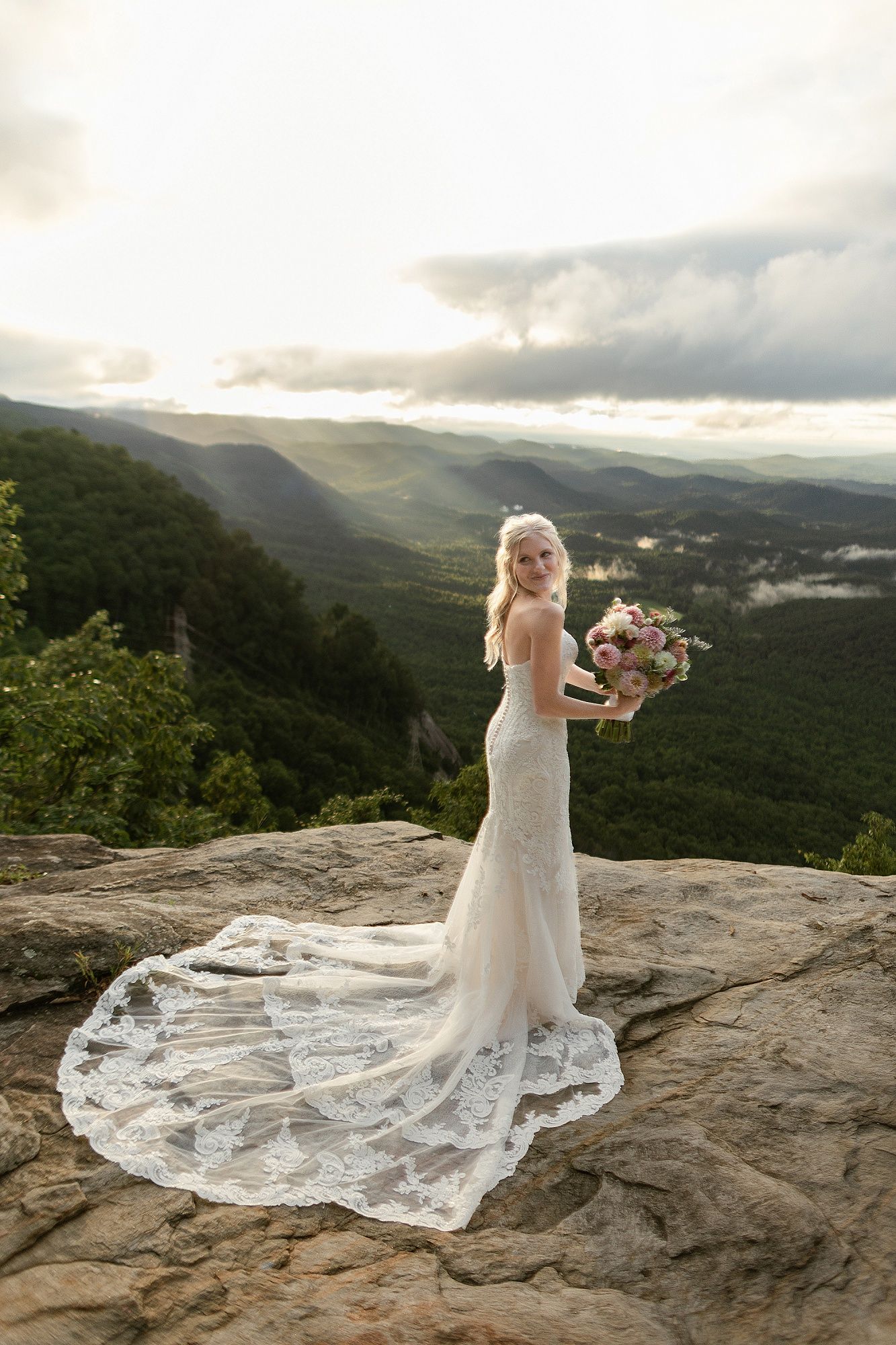 Bride in lace gown with long train holding bouquet on rocky mountain overlook at sunset.