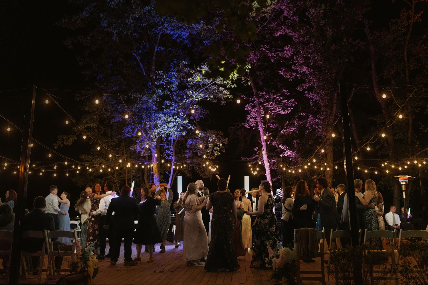 Outdoor evening wedding reception with string lights and colorful tree uplighting in purple, blue, and green.