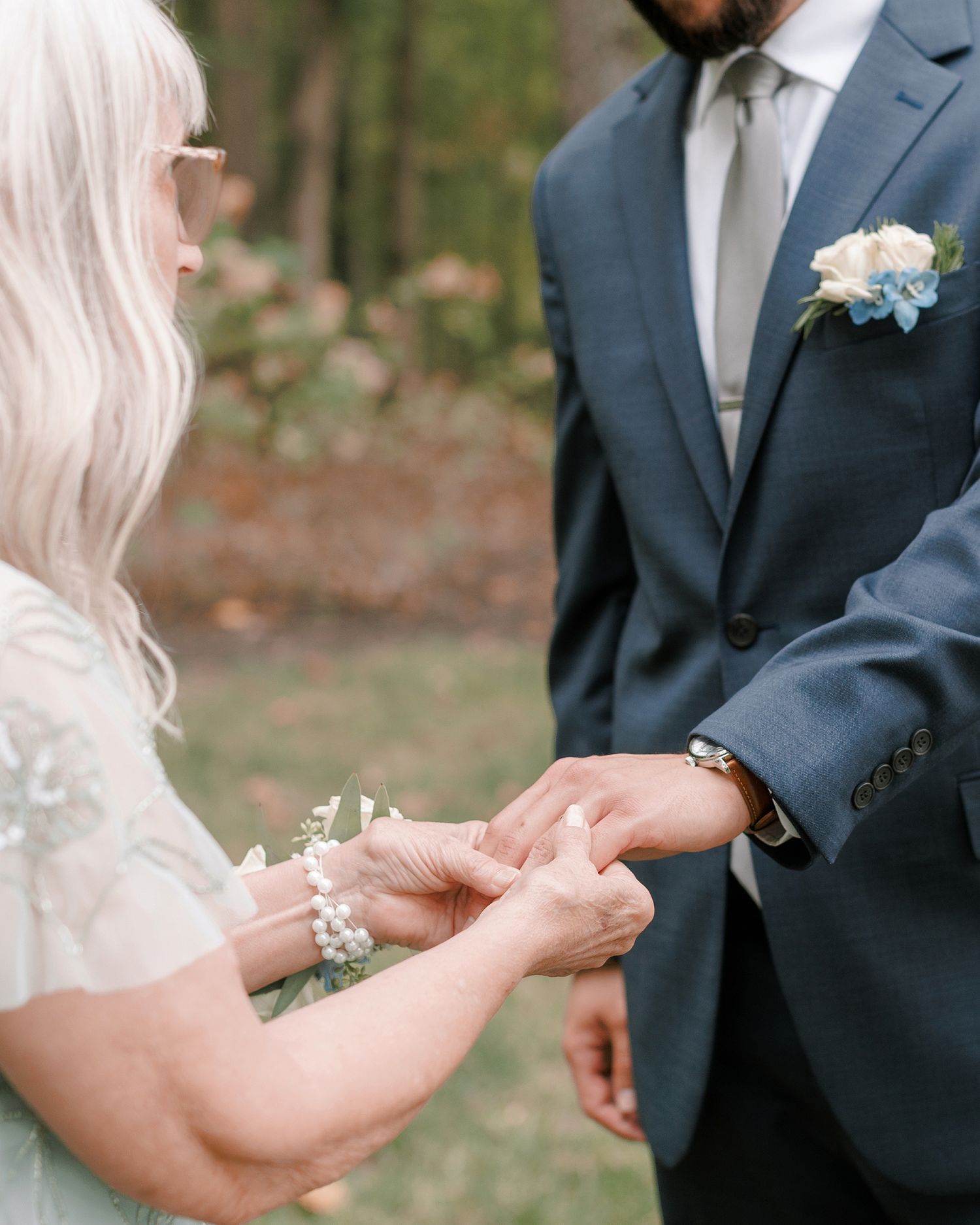Bride placing wedding ring on groom's finger during outdoor ceremony, groom wearing navy suit with floral boutonniere.