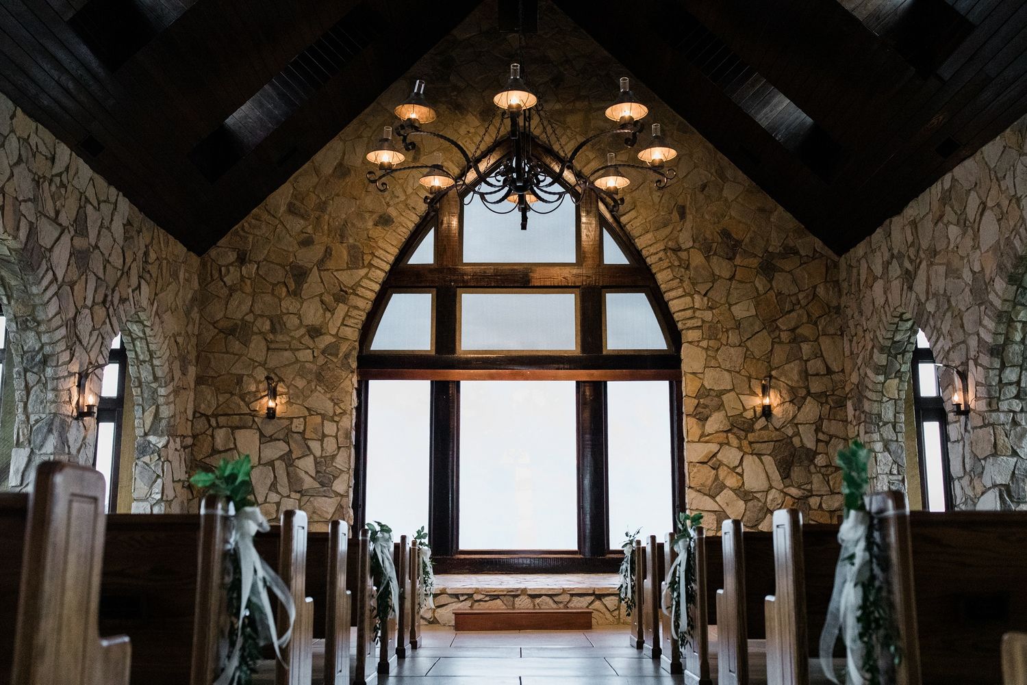 Rustic stone chapel interior with wooden pews, chandelier, and arched window above the altar.