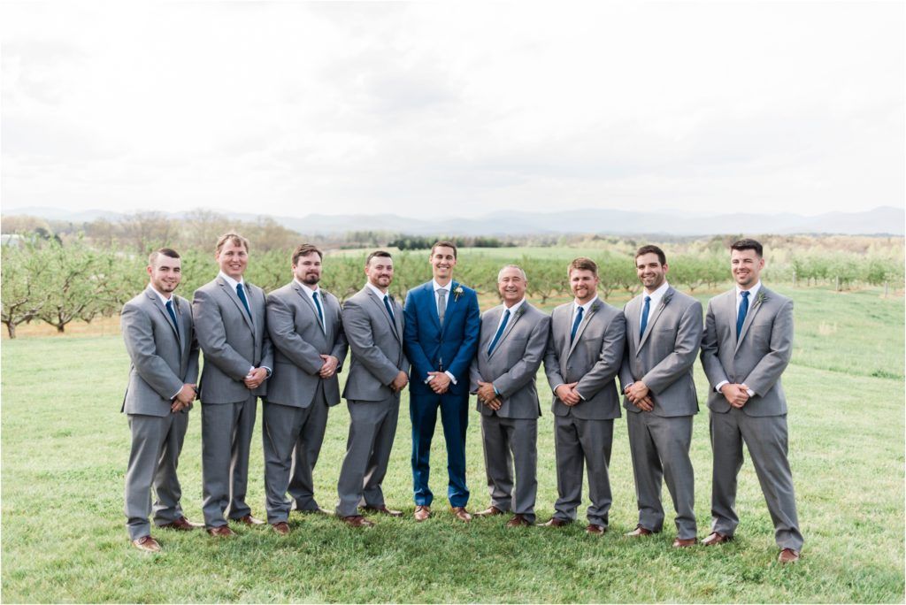 Groomsmen in grey suits with navy-suited groom standing in grassy field.