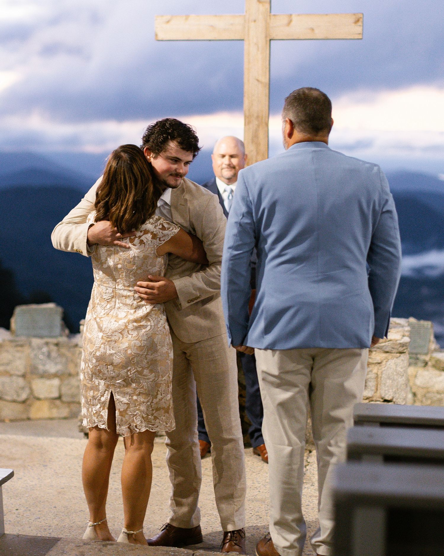 A couple embraces at a scenic mountaintop ceremony with a large wooden cross and an officiant overlooking the landscape.