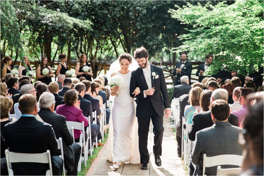 A wedding ceremony takes place outdoors with guests seated in white chairs along a tree-lined pathway.