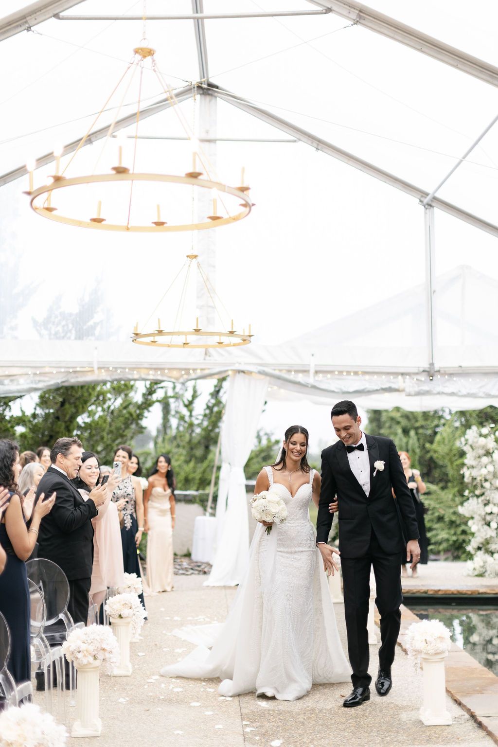 Wedding ceremony exit under a white tent with elegant chandeliers as guests applaud.