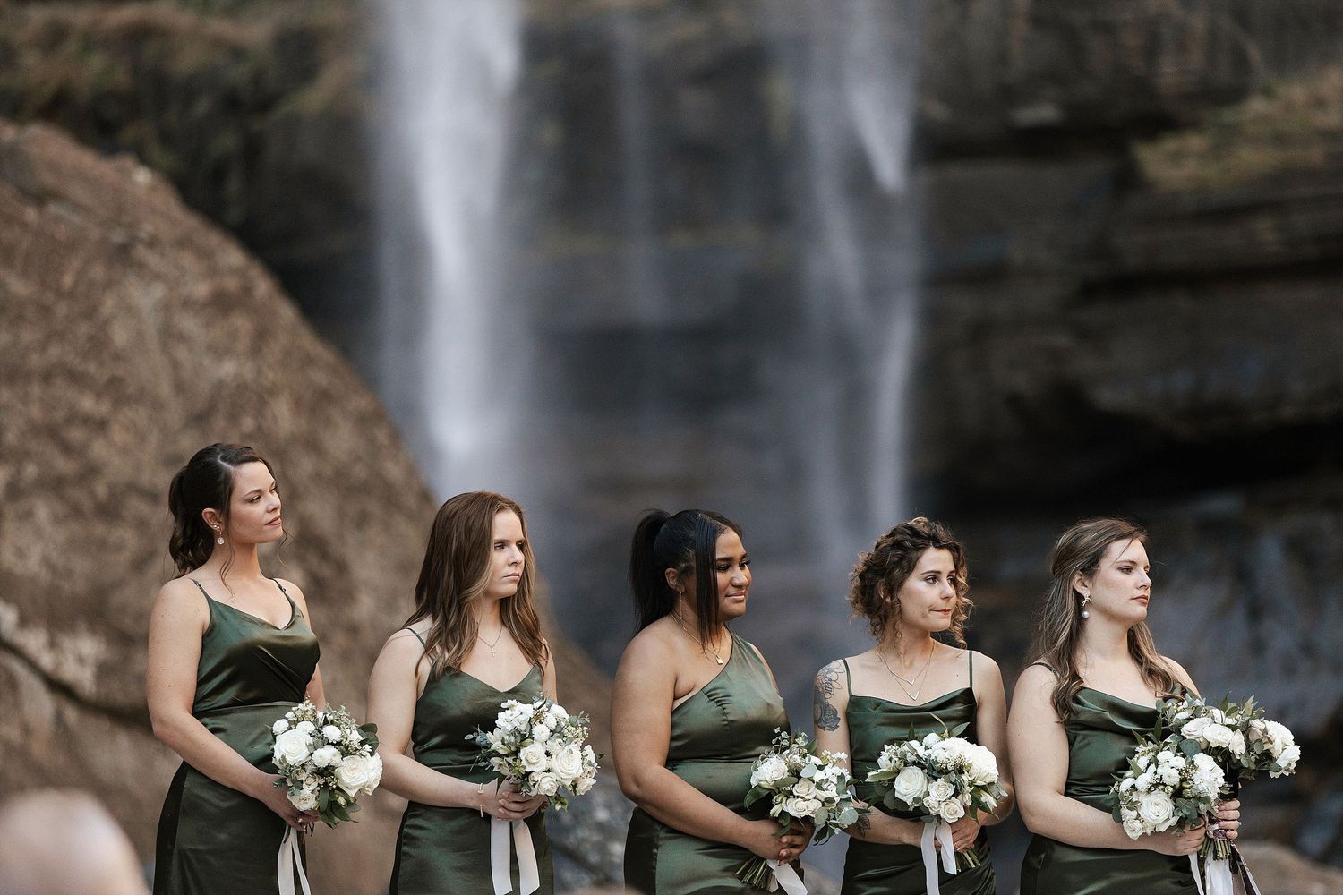 Five bridesmaids in sage green dresses stand in a line holding white bouquets as a waterfall cascades behind them.