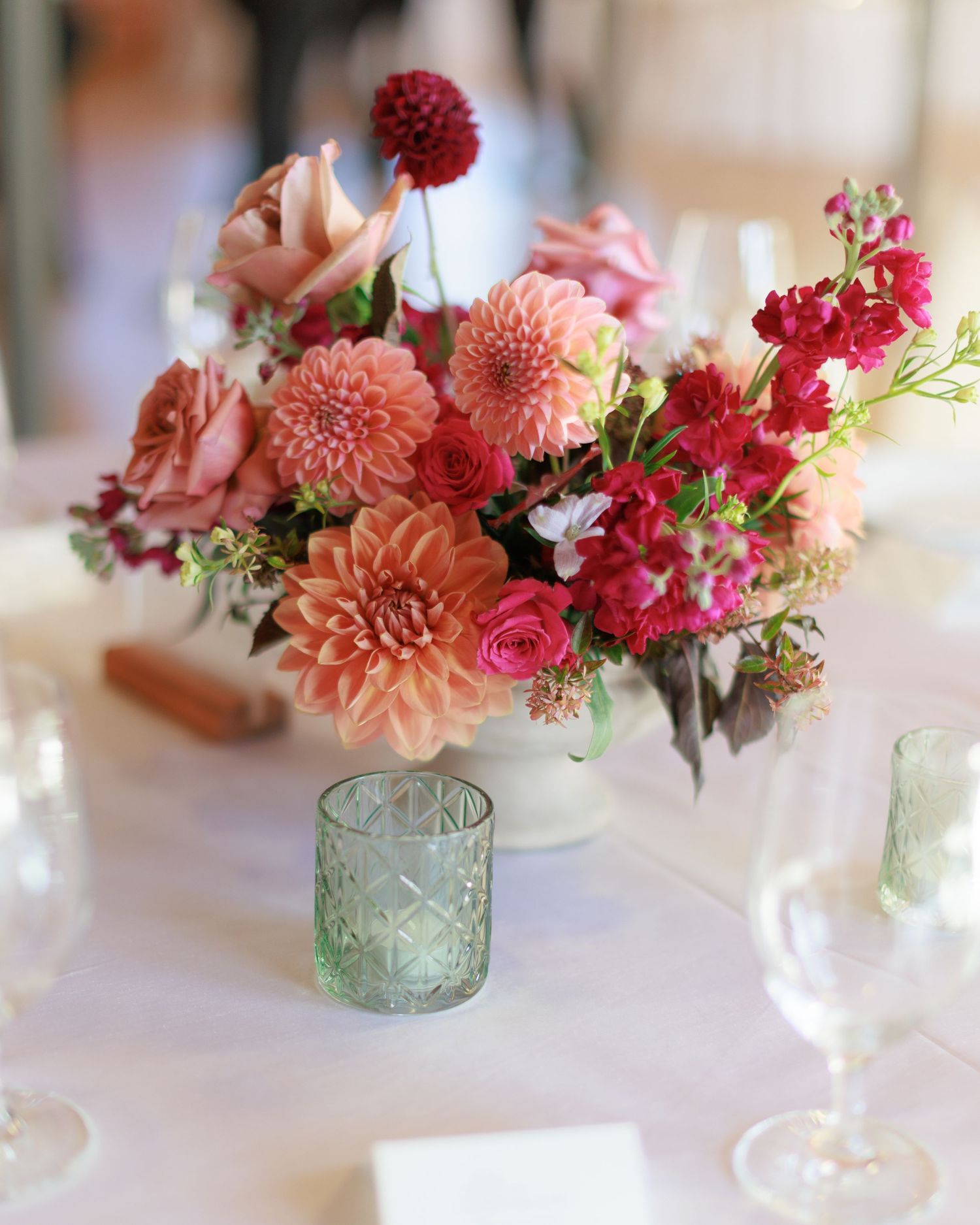 Pink and coral dahlia wedding centerpiece with roses in white vase, green glass votive candle holder on white tablecloth.