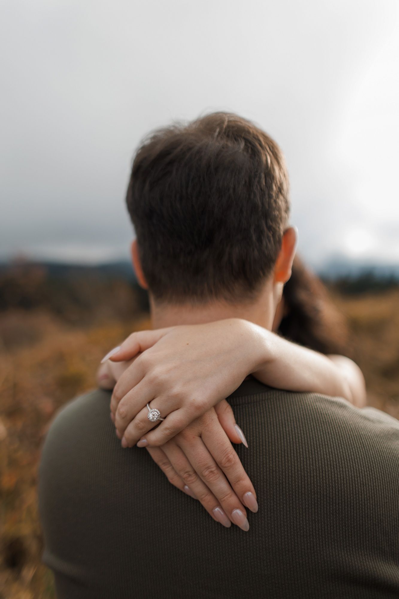 A close-up of hands embracing with an engagement ring visible against an olive green sweater in an outdoor setting.