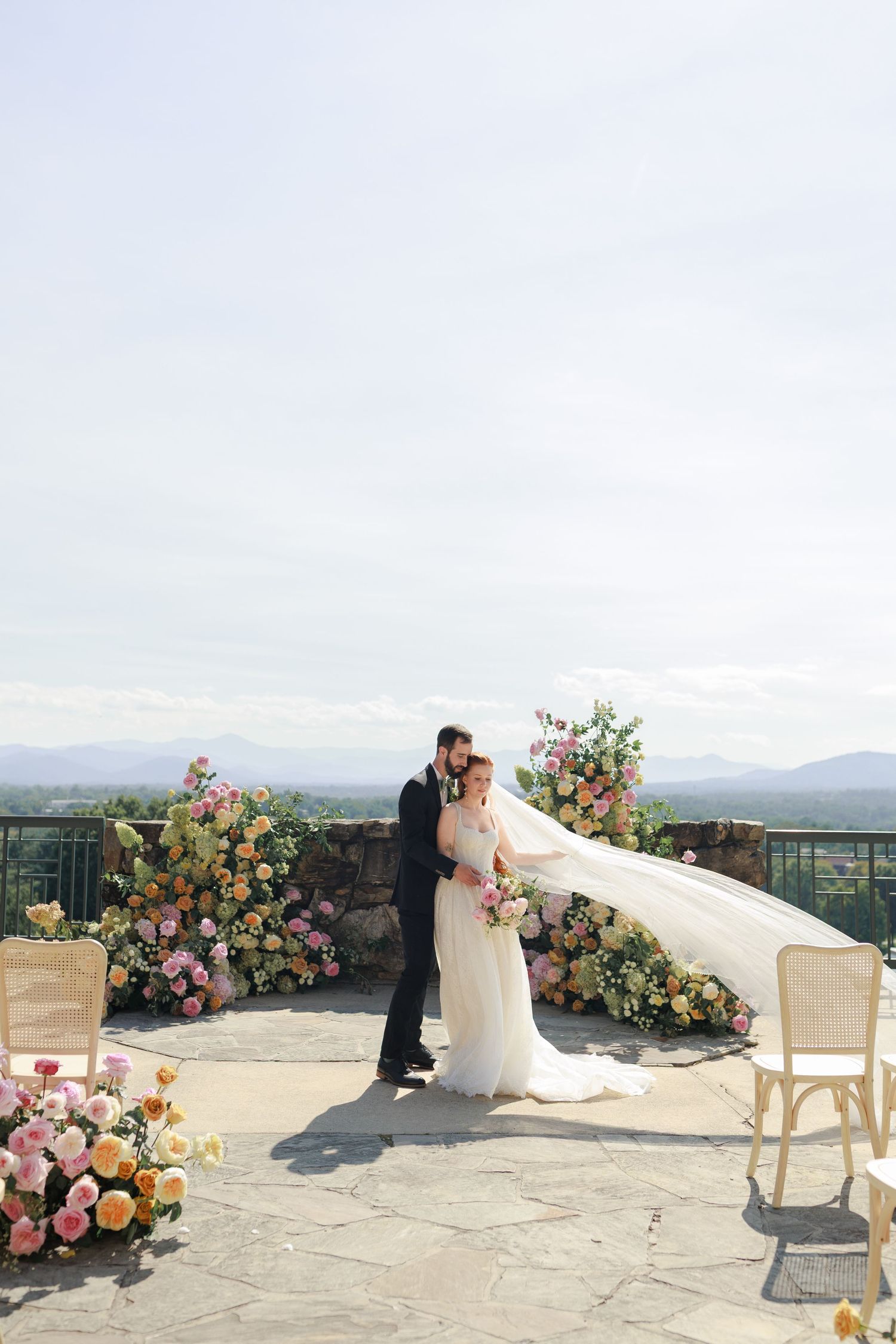 Bride and groom embrace at outdoor wedding ceremony with colorful floral arrangements and scenic mountain views.