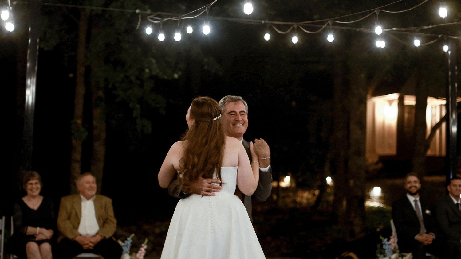 Bride and groom share first dance outdoors at night under string lights with wedding guests seated in background.