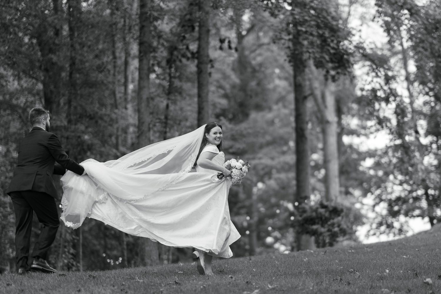 Black and white photo of a bride and groom walking outdoors, her veil flowing in the wind among tall trees.