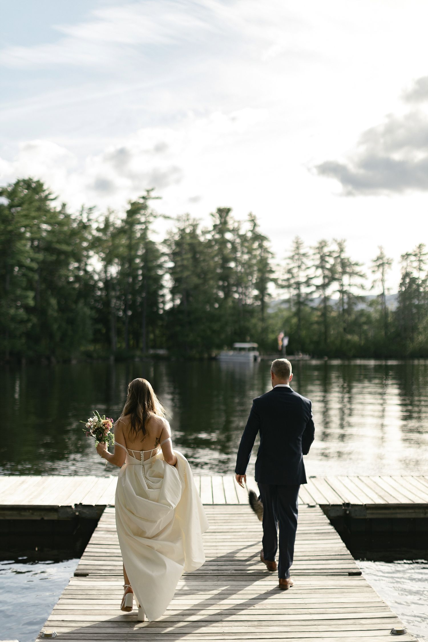 A couple walks along a wooden dock by a serene lake after their wedding ceremony.