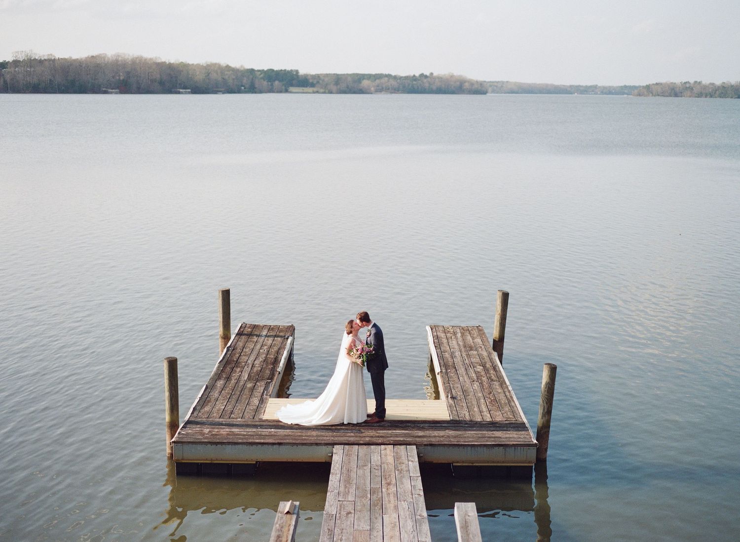 A couple stands together on a wooden dock over a calm lake on a misty day.