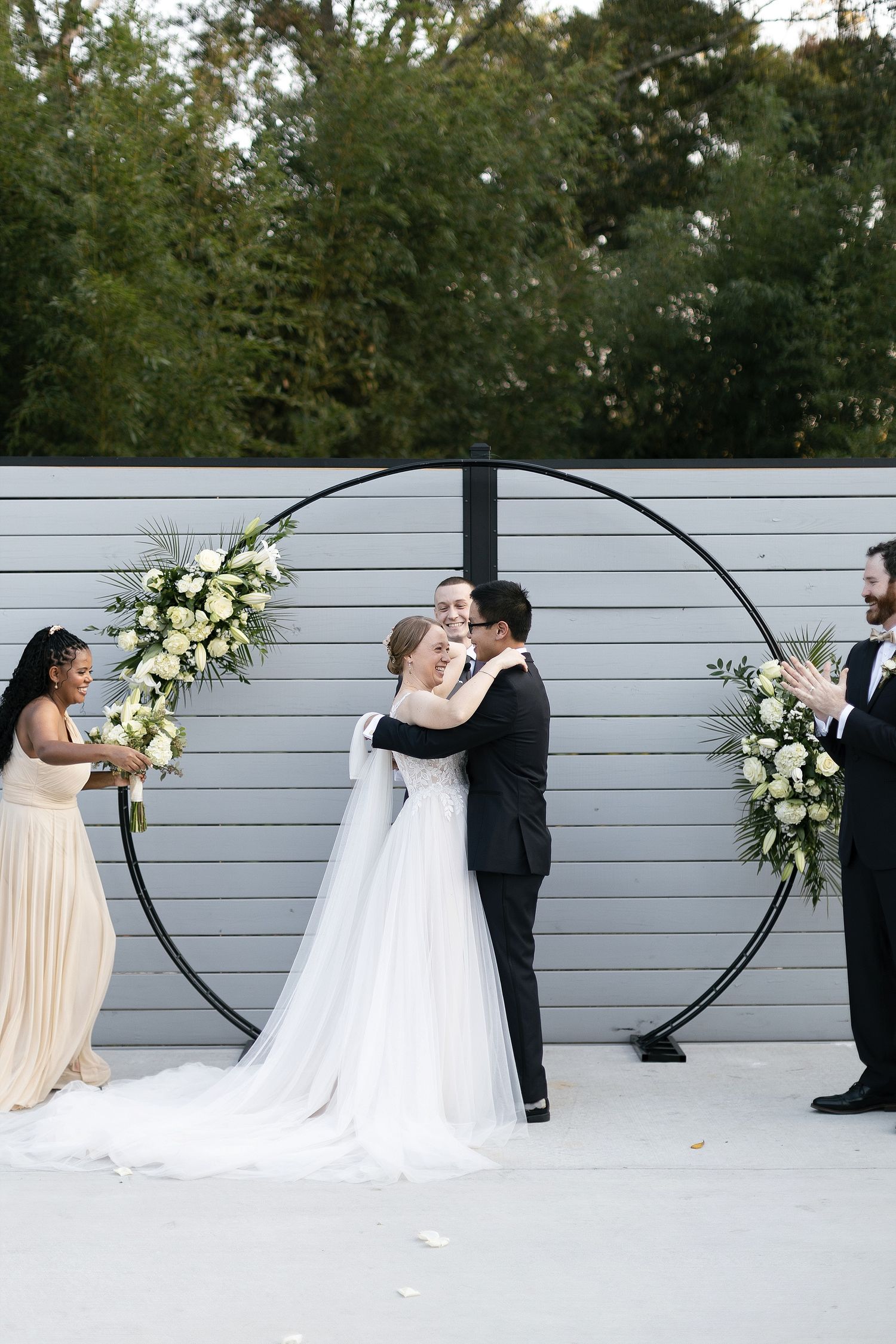 Minimalist wedding ceremony setup with a gray wall, black circular arch, and white floral arrangements.