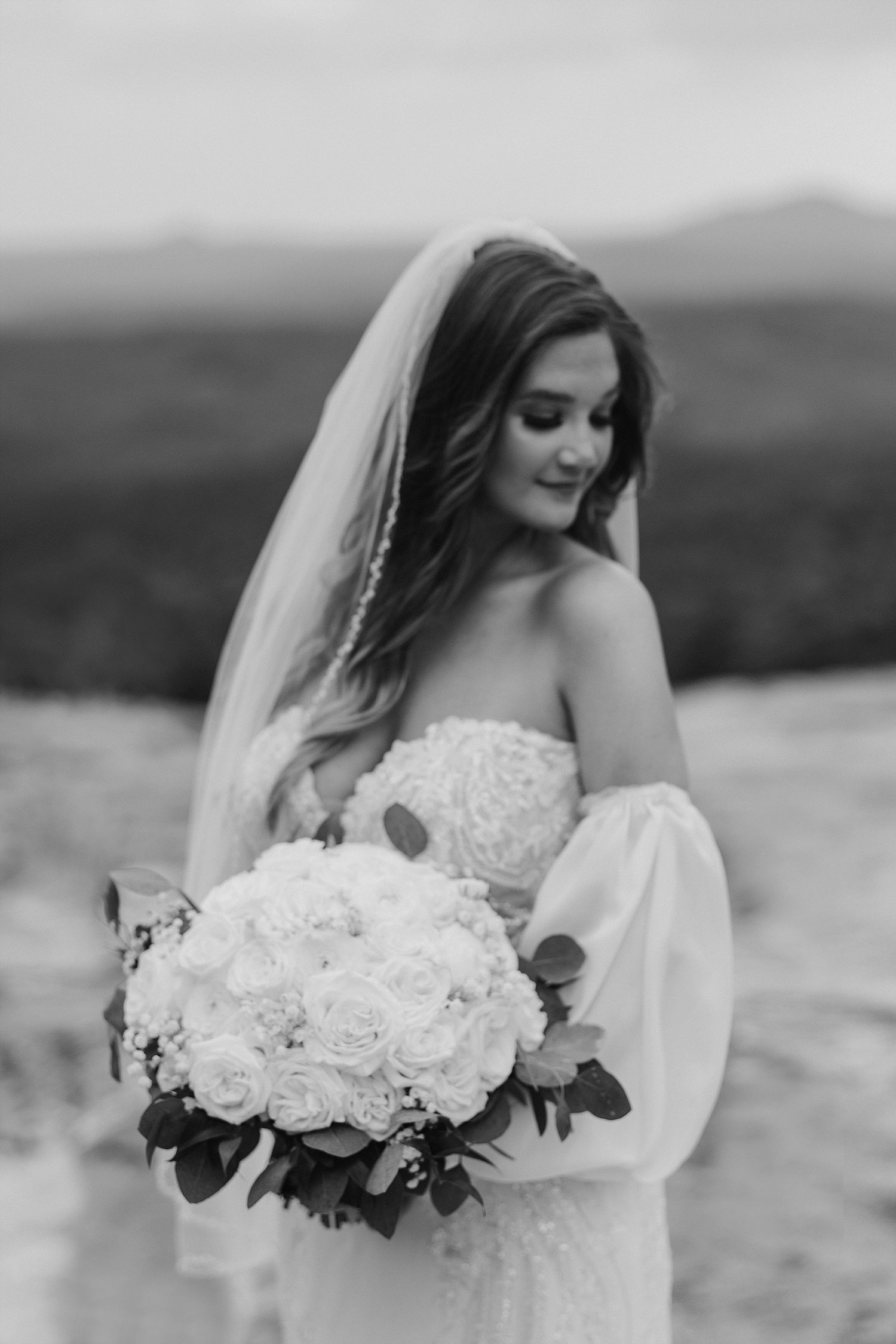 A black and white portrait of a bride holding white bouquet against mountain landscape.