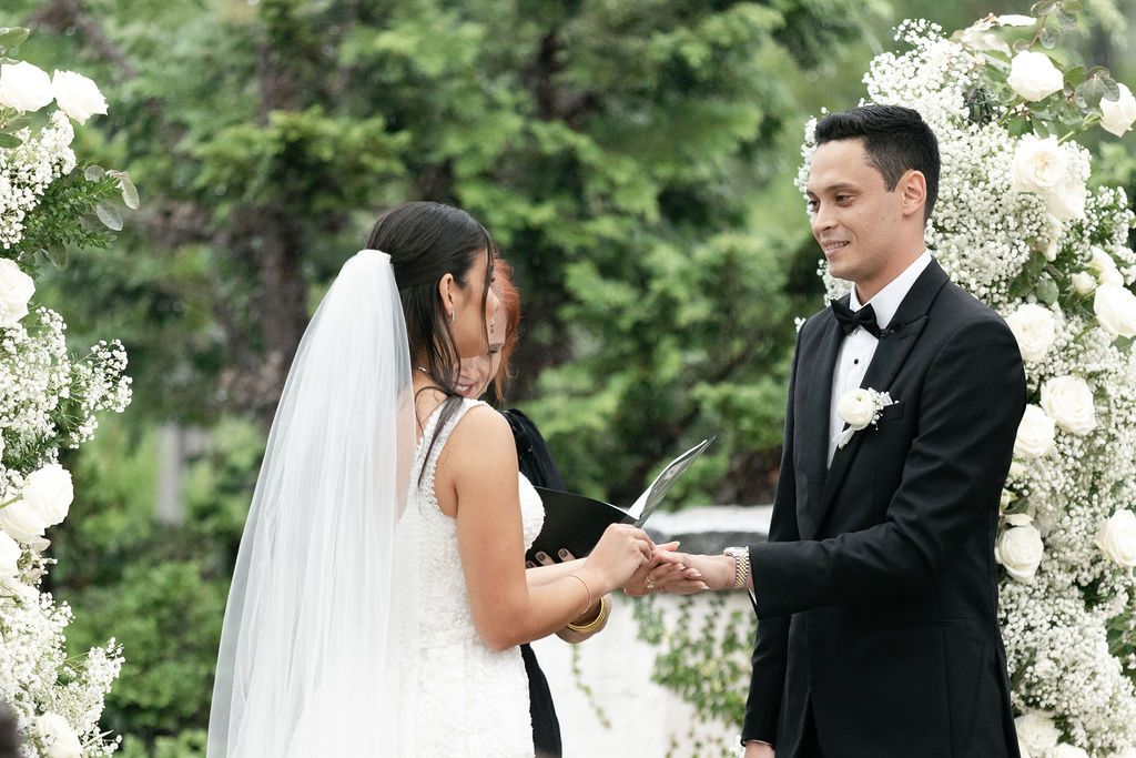 Exchange of rings during outdoor wedding ceremony with white floral arch.