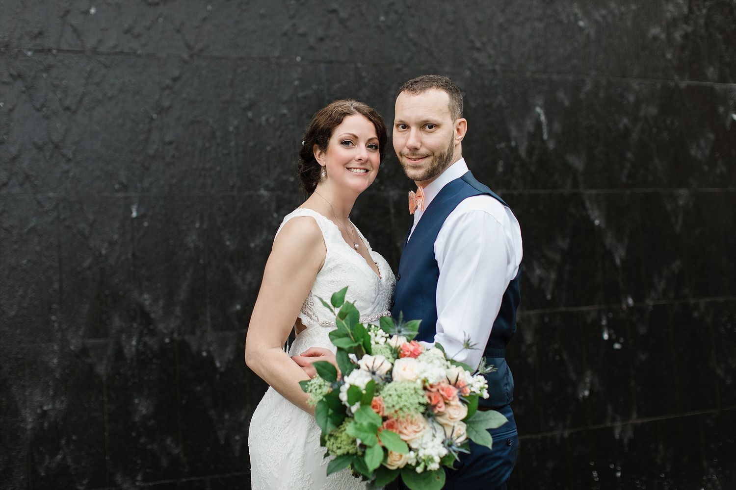 A couple poses against a dark textured wall with a pastel floral bouquet.