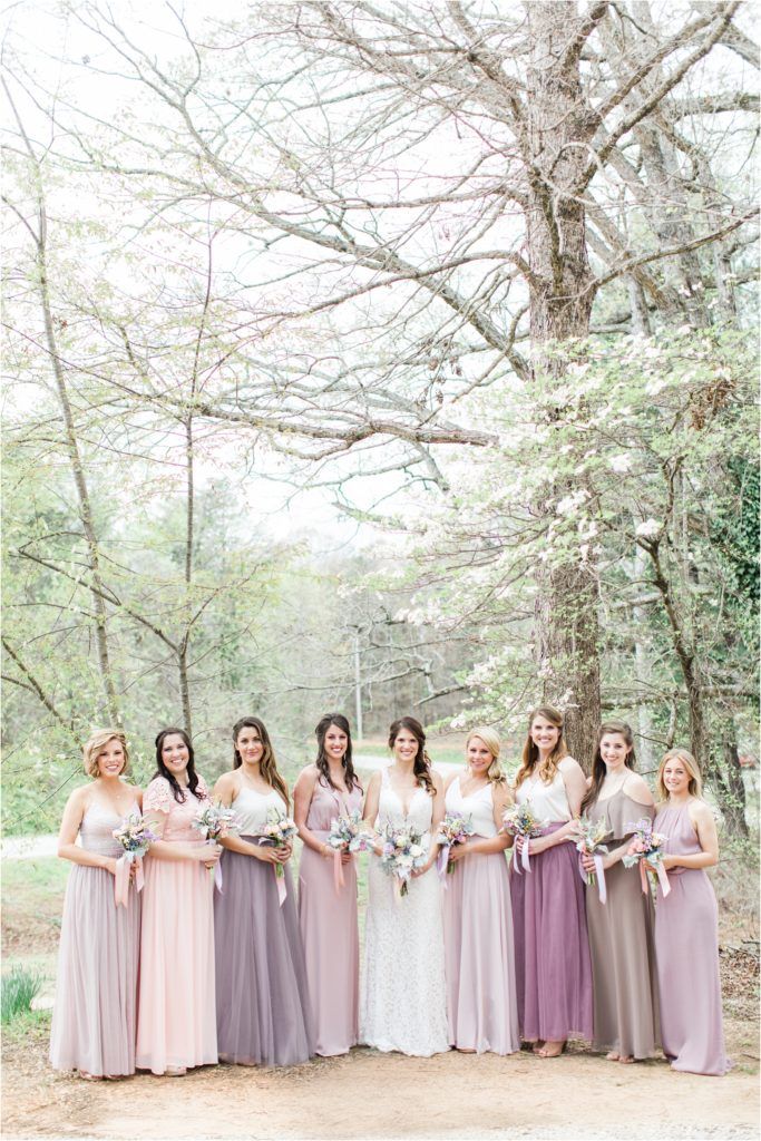 A wedding party in pastel purple and pink dresses standing together in a wooded setting.