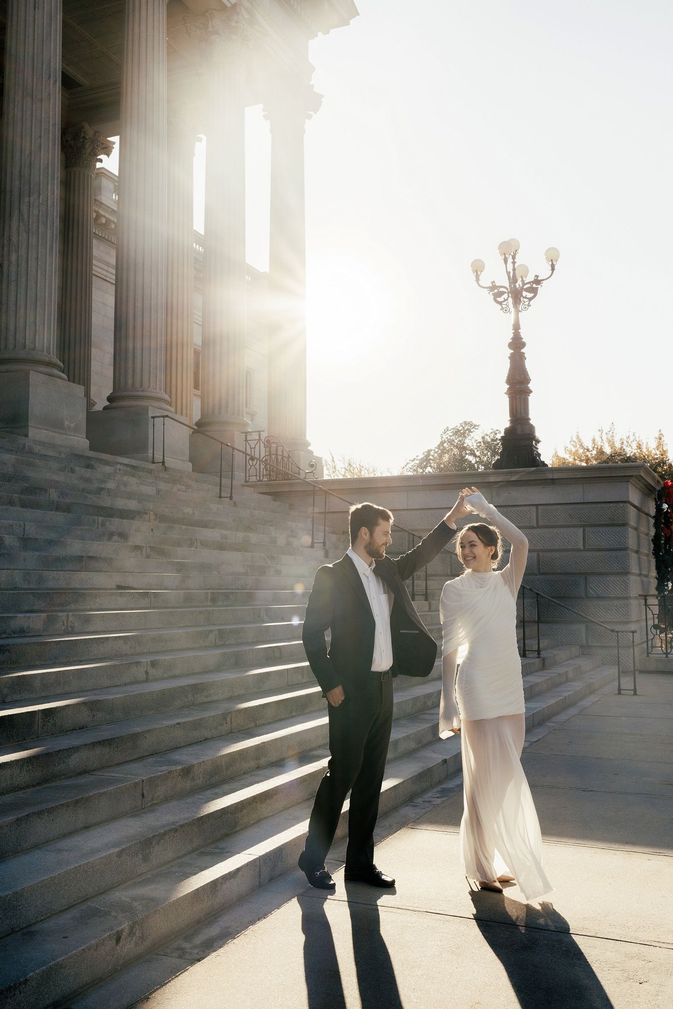 Couple dancing together on grand stone steps with dramatic sunlight streaming through classical columns in background.