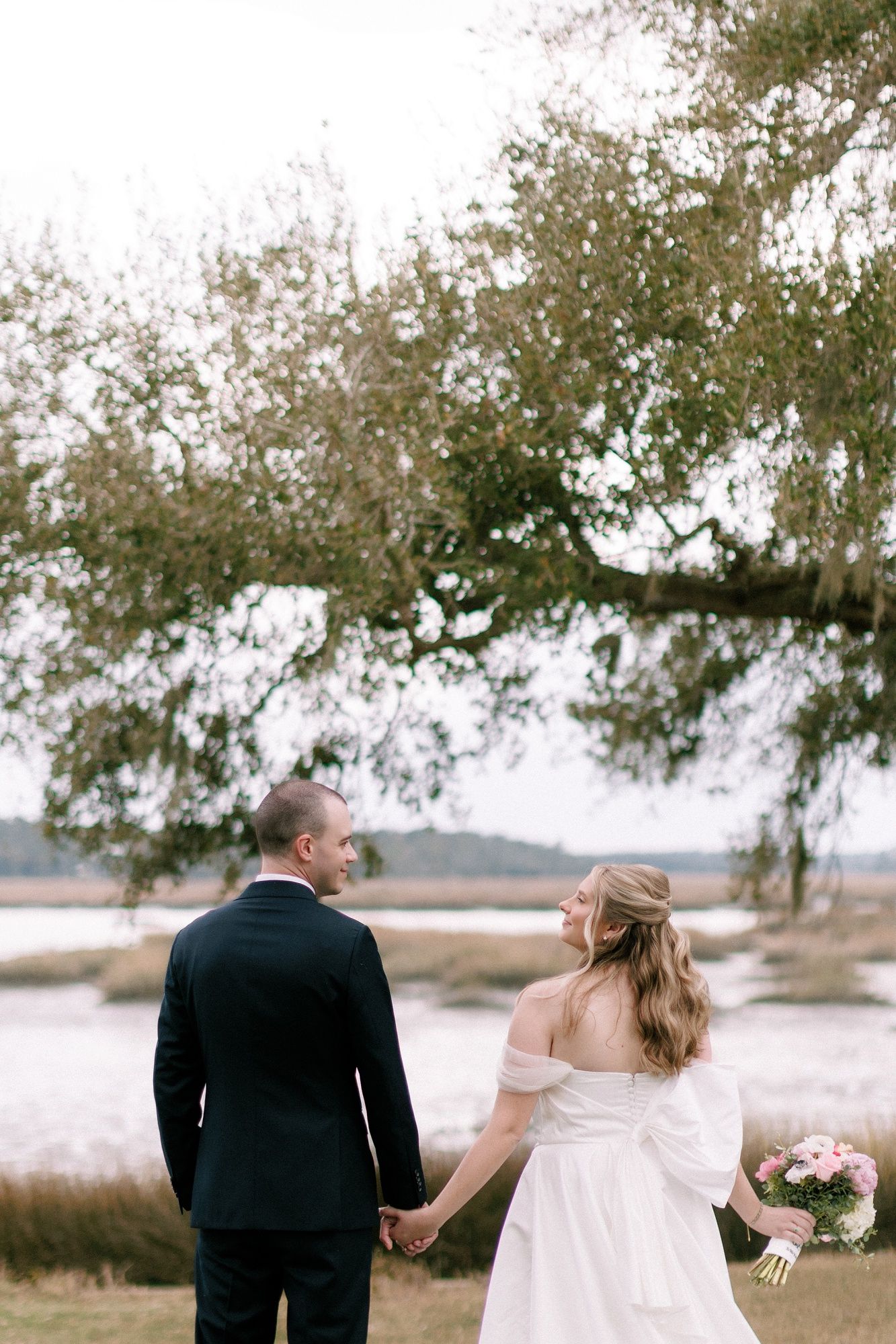 A couple in wedding attire walks together under oak trees draped with Spanish moss during their outdoor ceremony.