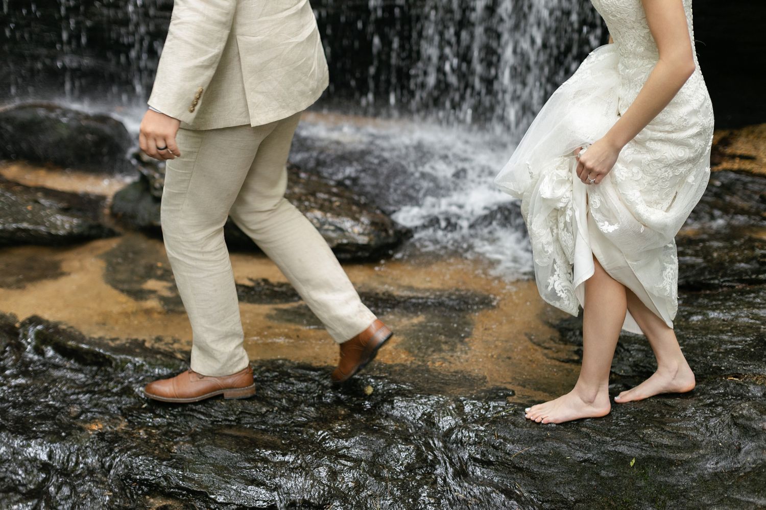 Two people in casual attire wade barefoot across a rocky stream with a cascading waterfall in the background.